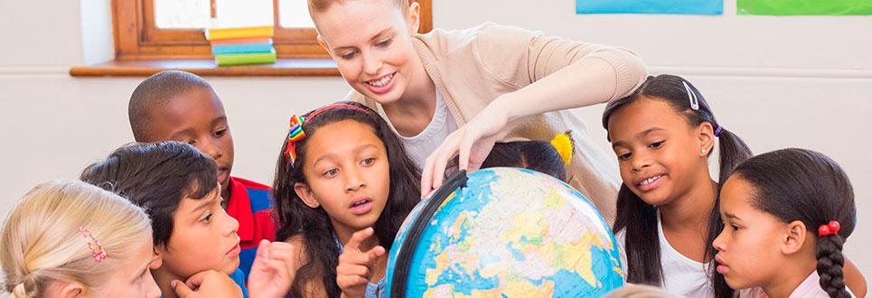 A group of younger students gathered around a globe pointing at various places, while the teacher points at a specific spot on the globe.